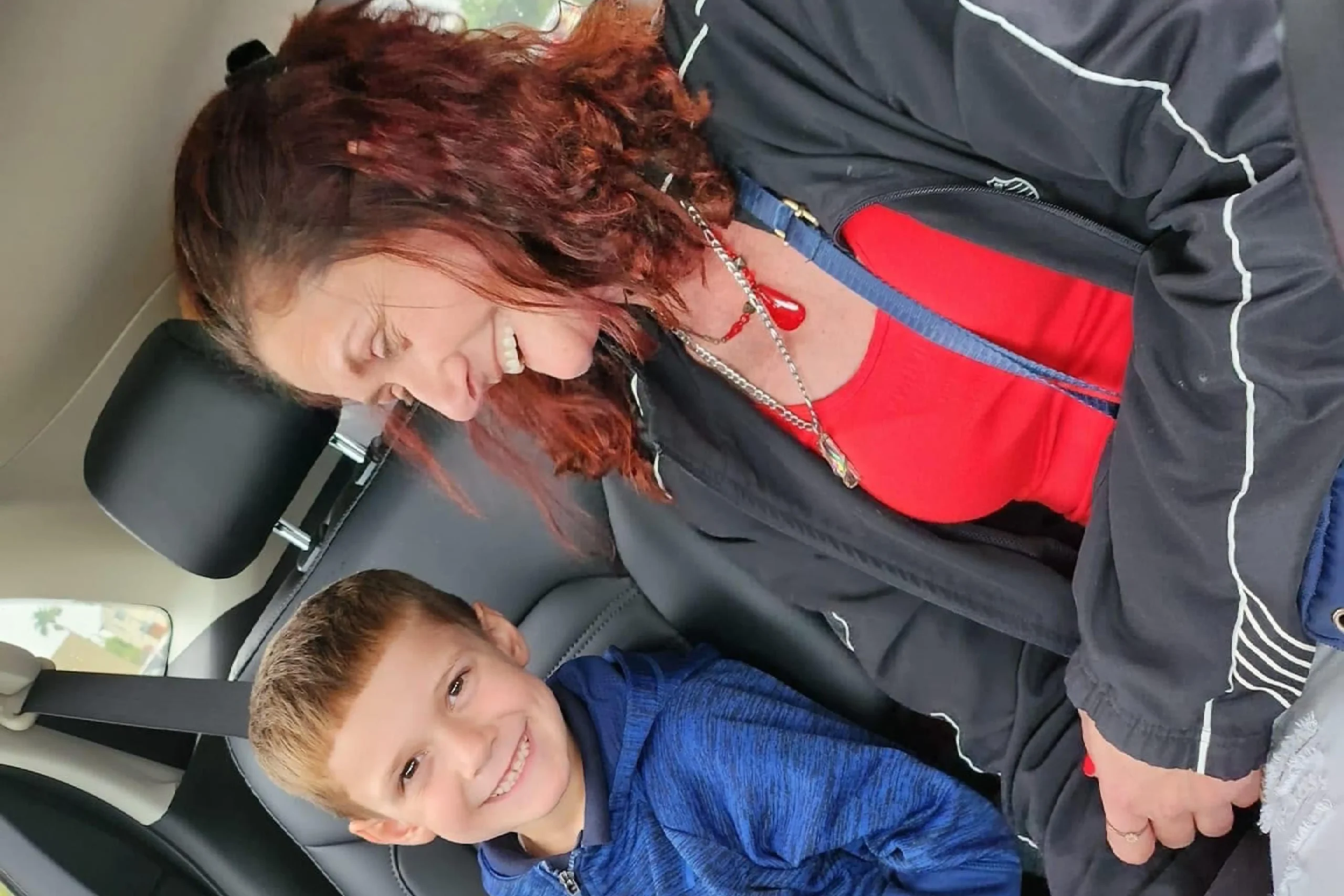 A person with curly hair wears a black jacket over a red shirt, seated in a car beside a child in a blue hoodie, both engaged with a device